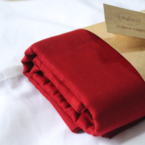 A folded piece of deep red turban fabric resting on a white surface, partially tucked under a kraft paper sleeve with a small tag reading CHARKHI TURBAN FABRIC.