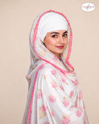 A woman wearing a white dupatta with pink floral prints and a pink border, paired with a white turban headwrap, set against a solid beige background. A 'CHARKHI' brand logo is visible in the top right corner.