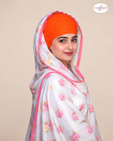 A woman wearing a bright orange turban and a white dupatta with pink floral prints and pink borders, photographed against a plain beige studio background. A Charkhi logo is visible in the top right corner.